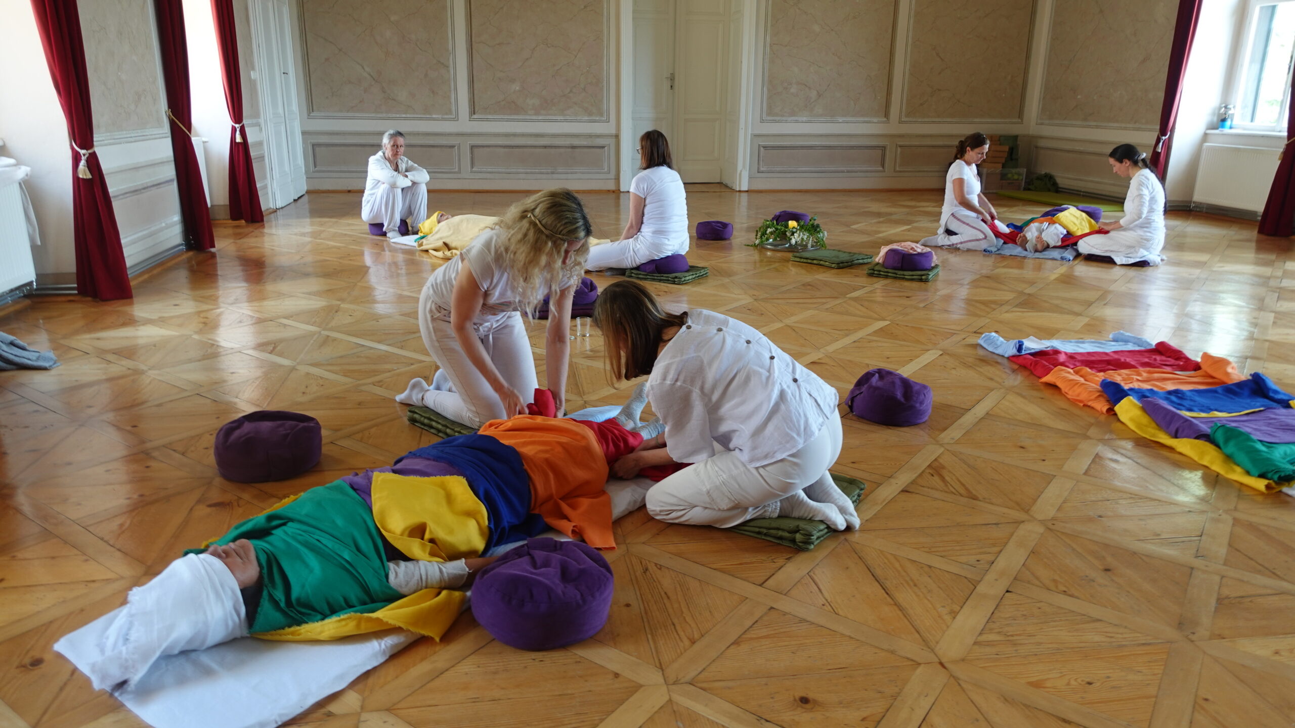 Qitreat Freiberg Gruppe vorne Praktizierende groß 2 Frauen in weißer Kleidung knien am Parkettboden des Festsaales Schloss Freiberg und machen eine Qitreat-Behandlung bei einer anderen Seminarteilnehmerin die in bunte Leinentücher gewickelt wird