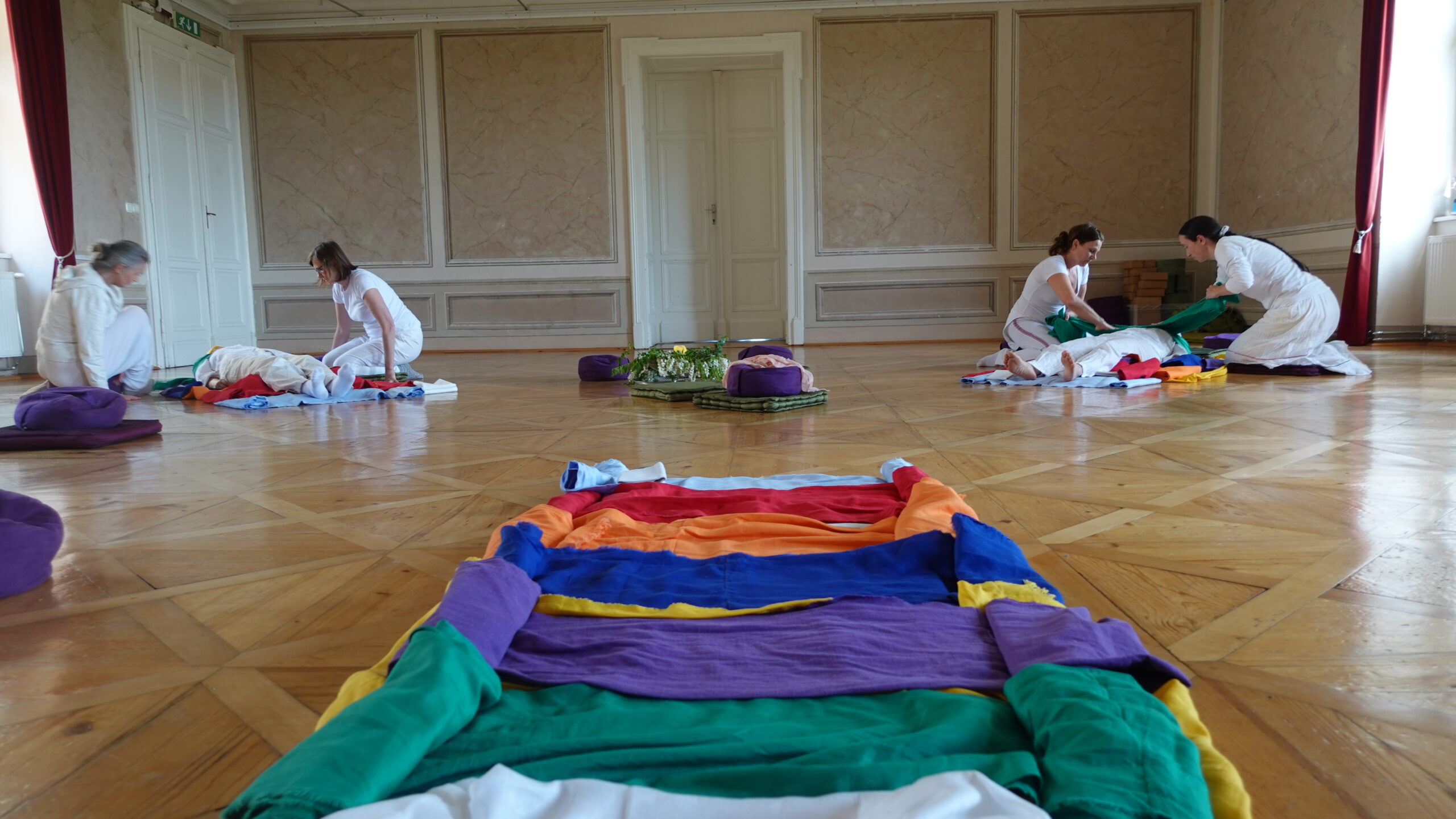 Qitreat Freiberg Gruppe vorne Tücher groß 3 Teams von je 2 Frauen in weißer Kleidung machen am Parkettboden des Festsaales im Schloss Freiberg machen Qitreat-Behandlungen im Rahmen eines Seminars bei je einer dritten Seminarteilnehmerin die in bunte Leinentücher gewickelt wird